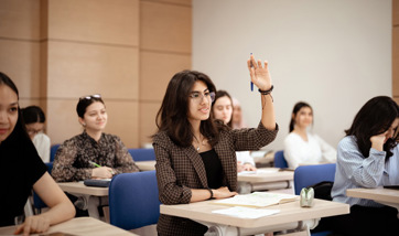 A young woman holds her hand up to ask a question, seated at an individual work desk in a classroom surrounded by other students.