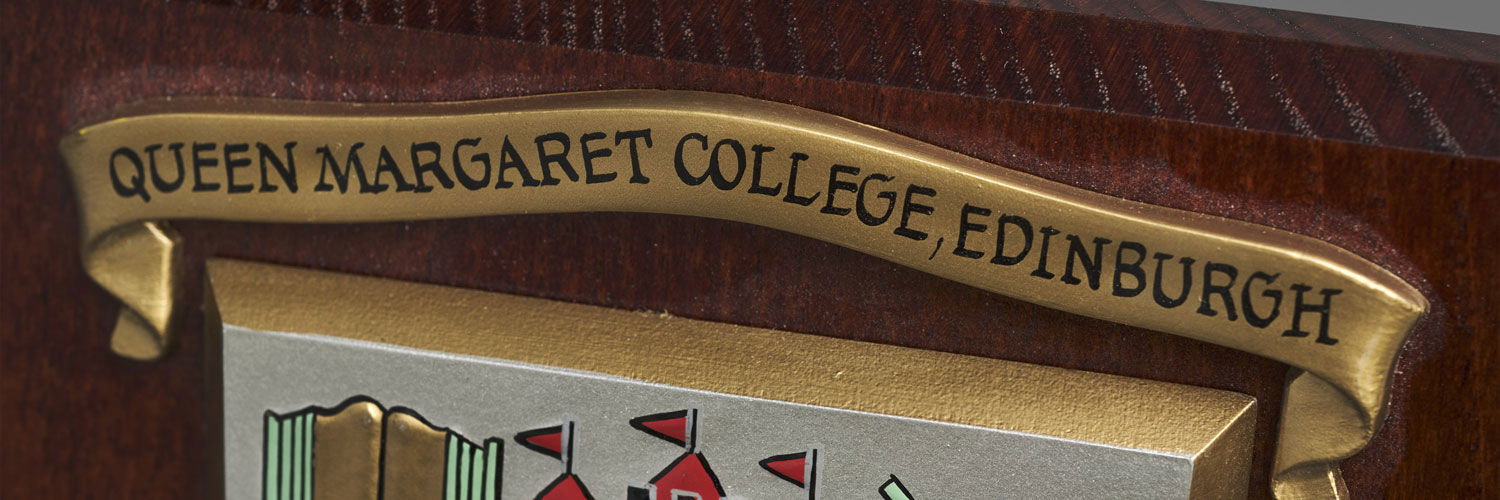 The Queen Margaret University College crest and motto, "with Head, Heart and Hand" affixed to a wooden shield.