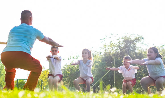 A teacher and a group of five children outside in a park performing squats.