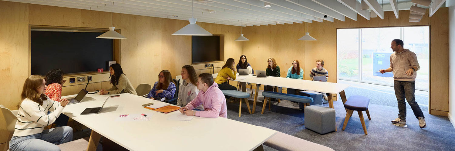 A teaching space within the library, there are two large tv screens, a whiteboard, and two long tables filled with students working on laptops.
