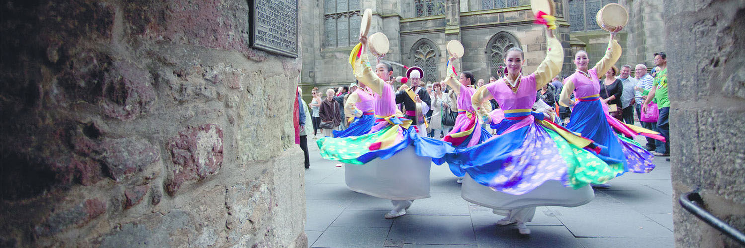 A group of dancers performing on the streets of Edinburgh in colourful dresses with a crowd of people watching them