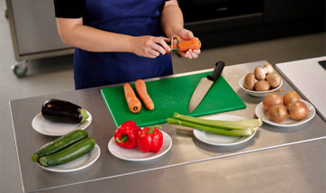 A person peels a carrot over a food preparation table laid with various vegetables.