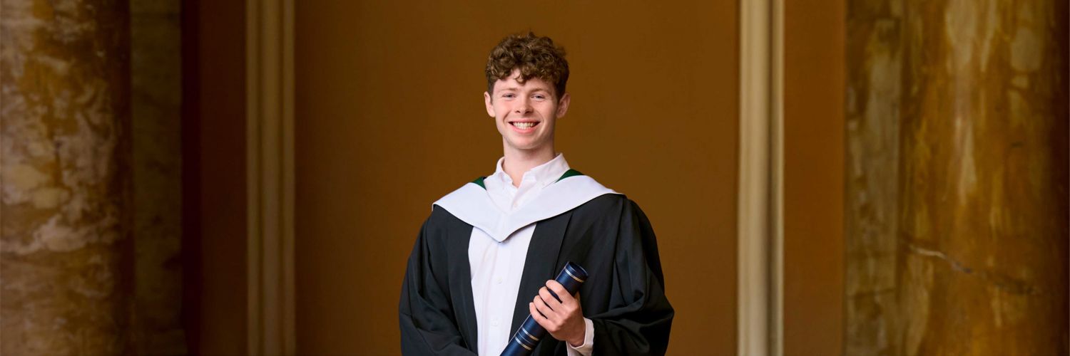 smiling young man wearing graduation robes and holding graduation scroll
