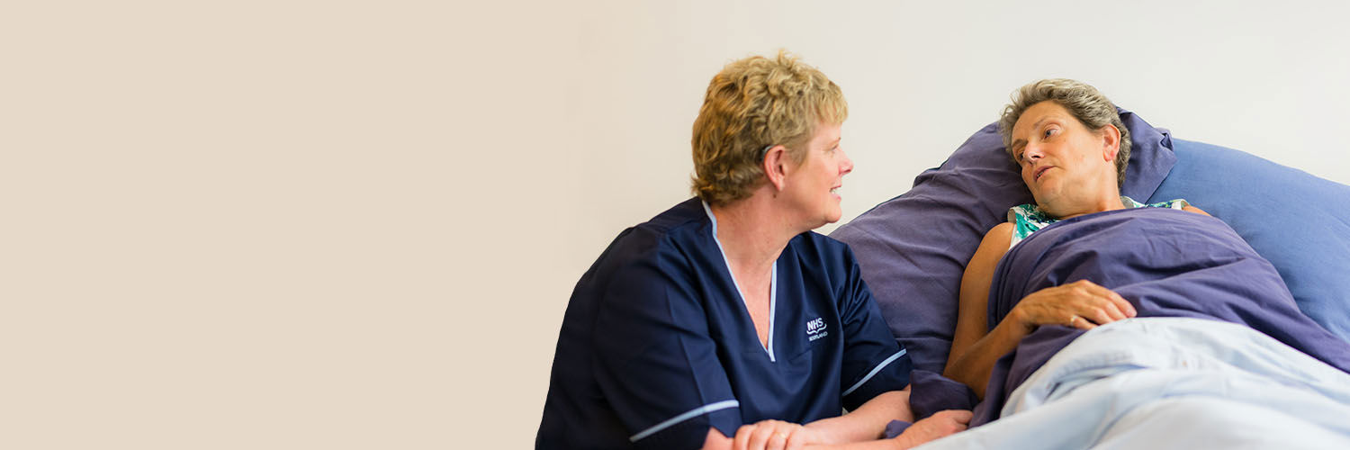 A palliative care nurse sitting by a patient's bed speaking to her