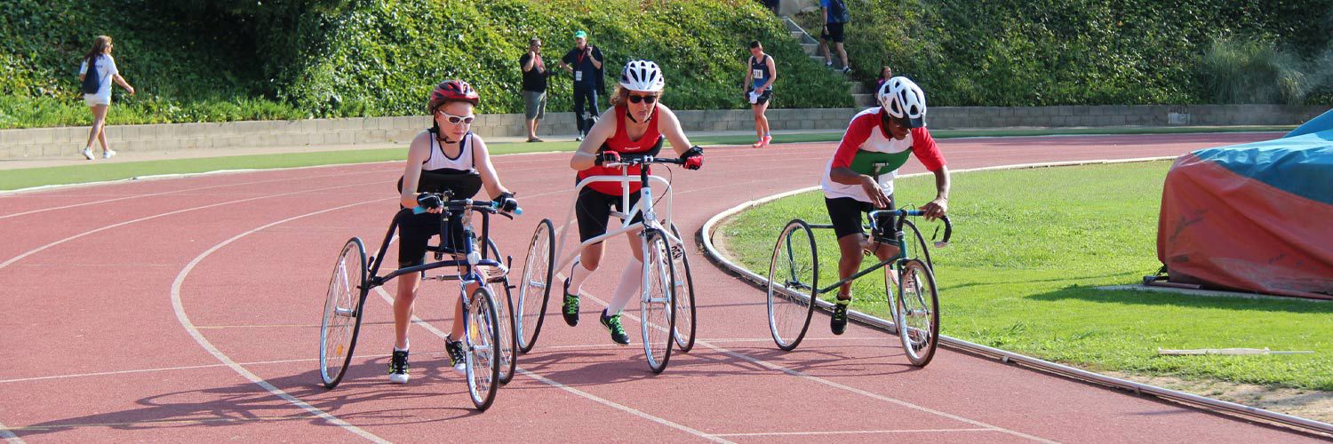 Three people using a three wheeled frame with a saddle, handlebars and a chest support (but no pedals) around a racetrack.