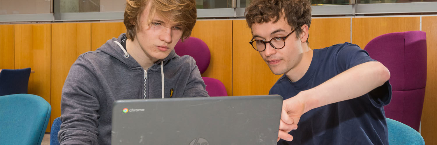 A young man tutoring another on a task involving a laptop.