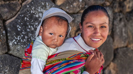 A young woman carries an infant on her back, tied securely with a brightly coloured fabric.