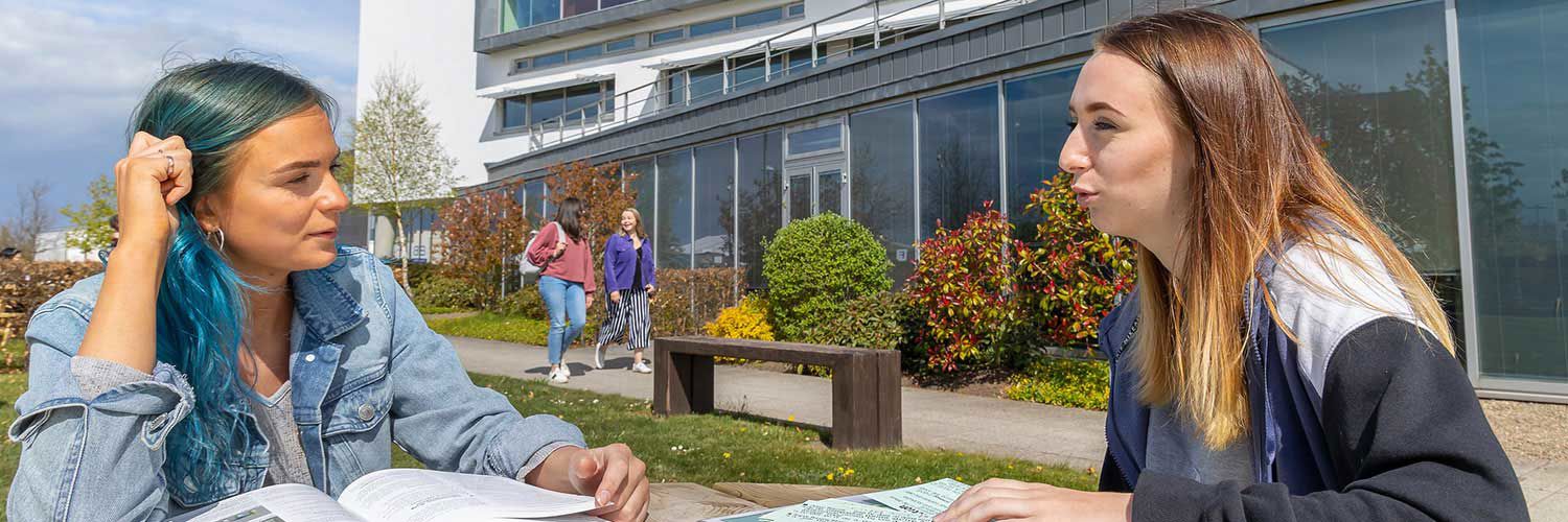 Two female students chatting over books in the outdoor space at QMU